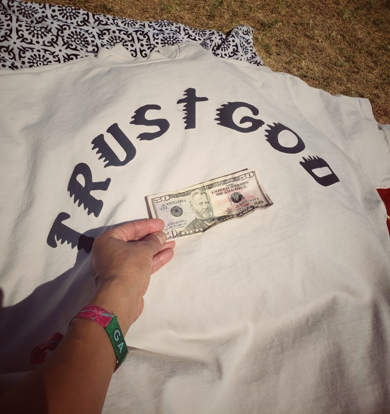 A fifty dollar bill in hand on top of a trust God T shirt