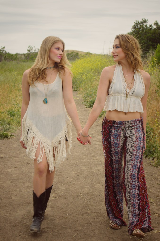 Two young women wearing boho style clothing holding hands while walking down a hiking path with yellow wildflowers