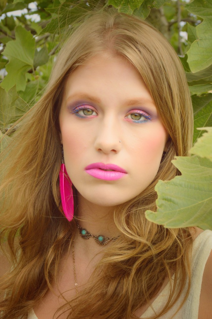 Portrait of a young woman standing underneath a tree surrounded by leaves wearing a pink feather earring.