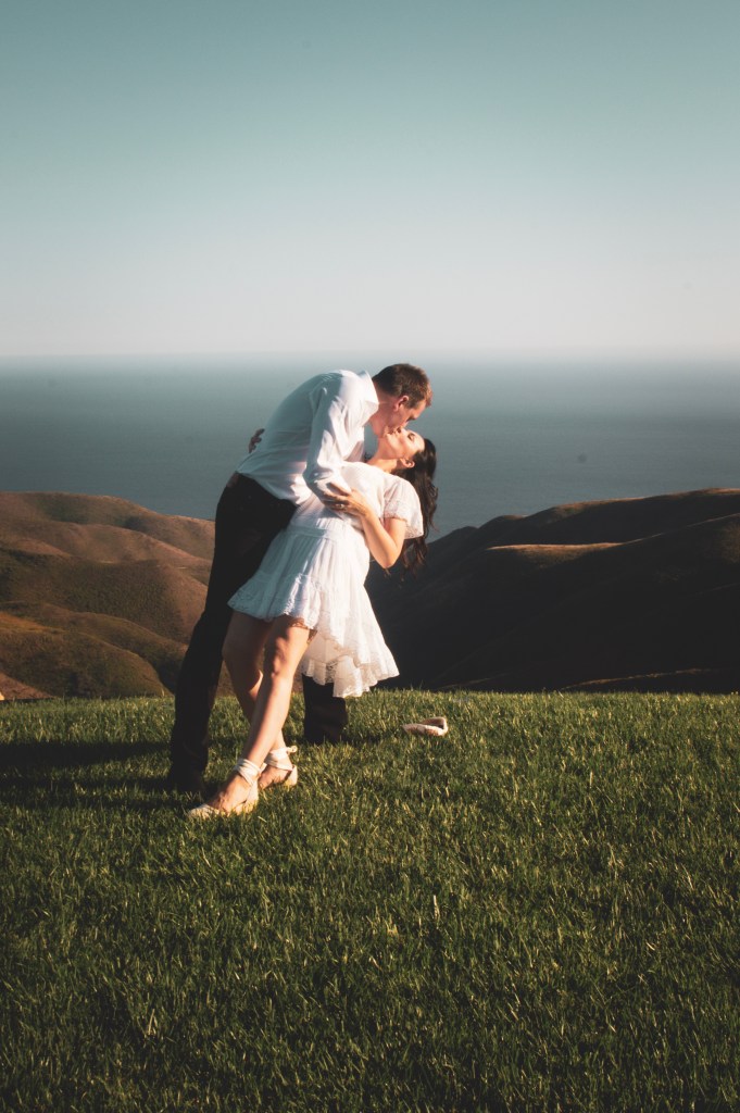 Man dipping a woman for a kiss on a mountainside overlooking the ocean