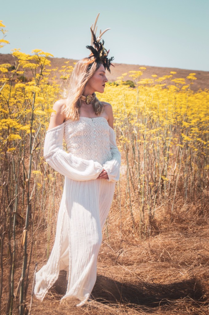 Blonde Woman standing in a mustard field in a white dress wearing a horn head piece