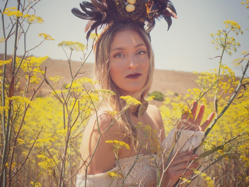 Blonde Woman standing in a mustard field in a white dress wearing a horn head piece