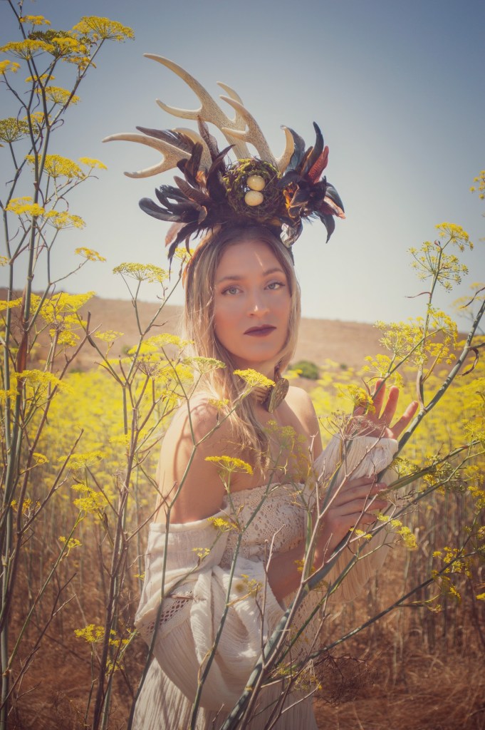 Blonde Woman standing in a mustard field in a white dress wearing a horn head piece