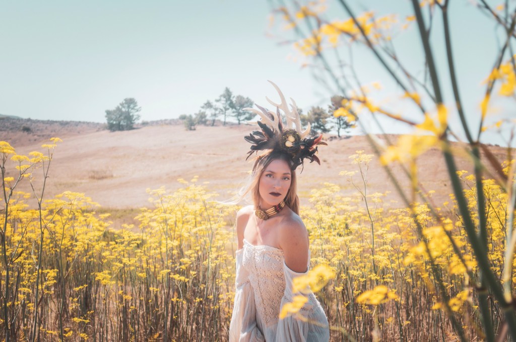 Woman standing in a mustard field in a white dress wearing a horn head piece